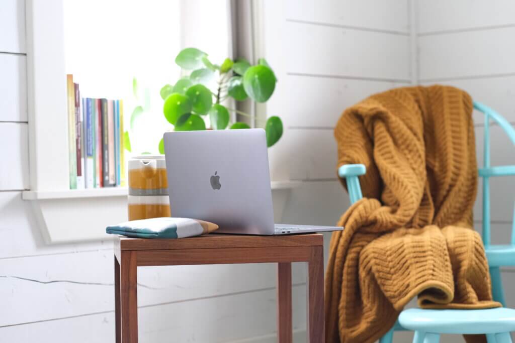 A laptop in a wooden table is placed in front of a blue chair with a knit blanket draped over it inside a brightly lit room.