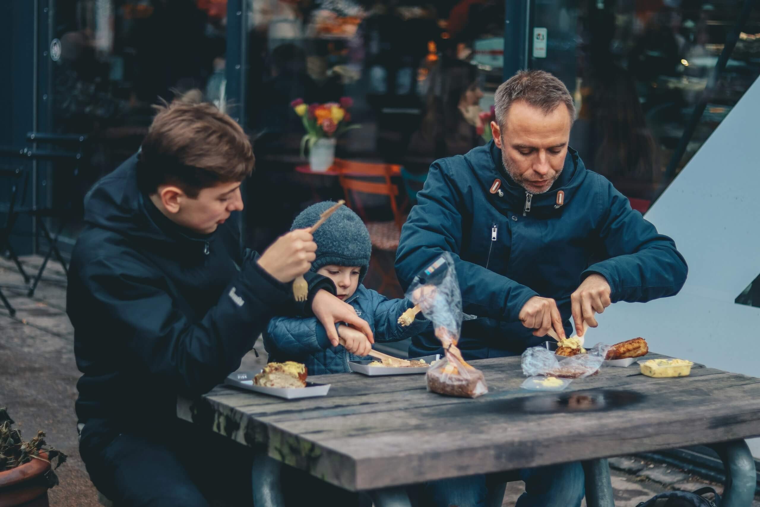 Three individuals (one adult, one teenager, one child) eating a meal at a table outside.